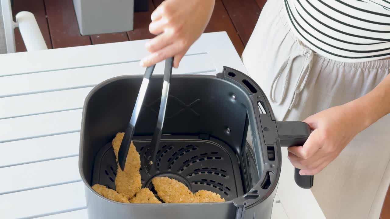 Person uses tongs to arrange crumbed chicken tenders in an air fryer basket on a white table, with bright natural lighting and steady overhead camera