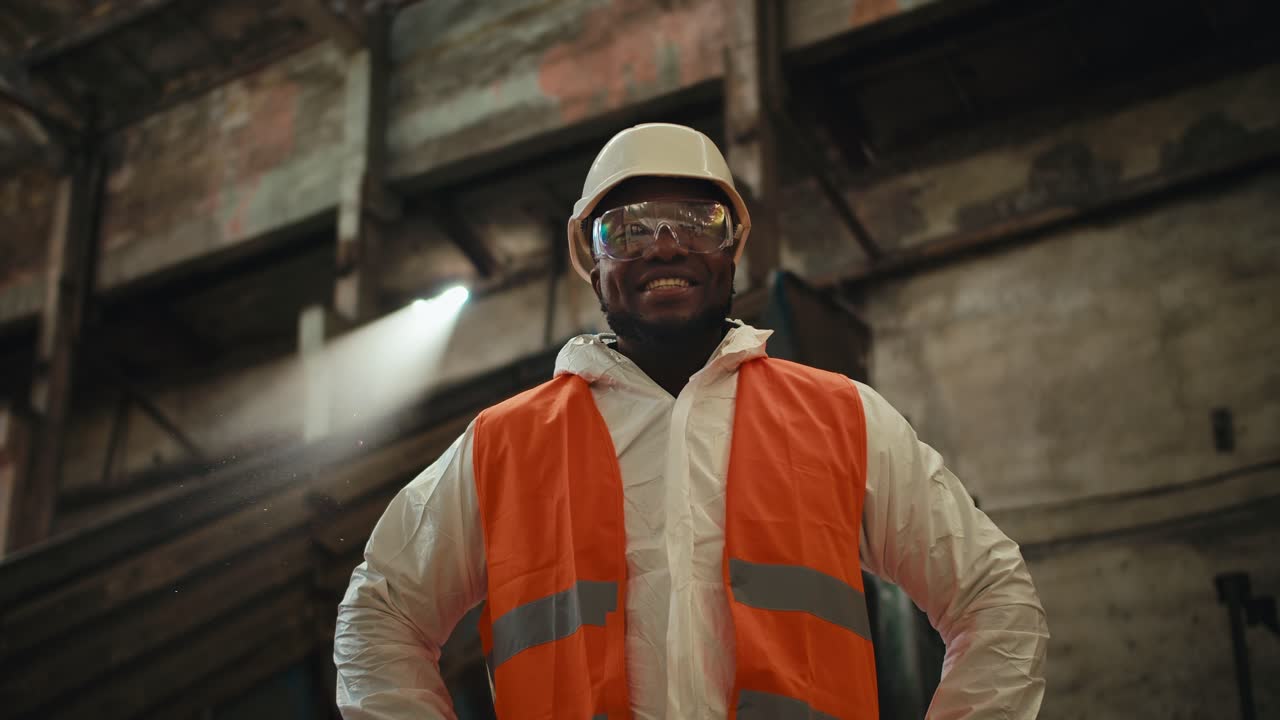 Close-up portrait of a happy man with Black skin in goggles and a white helmet in a special white uniform and an orange vest who stands in a huge factory near the conveyor belt
