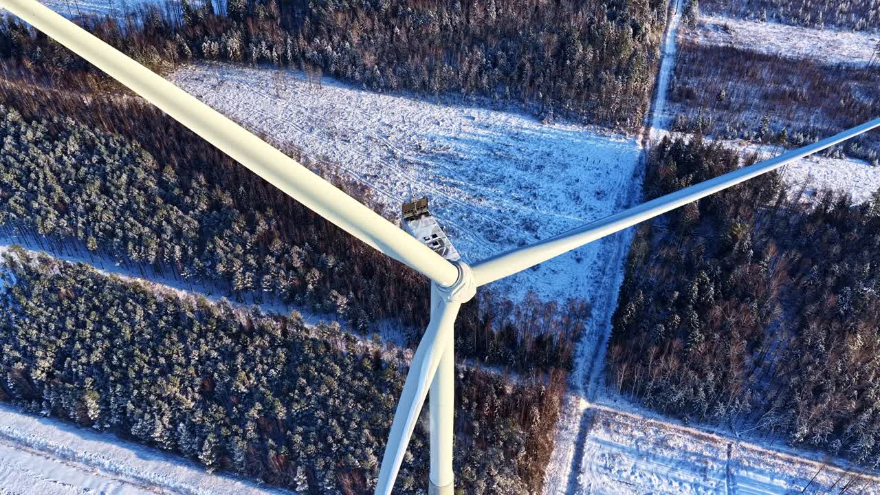 Sorvolo aereo inclinato verso il basso sopra una turbina eolica immobile, circondata da radure innevate nella foresta durante un bellissimo sorgere del sole invernale