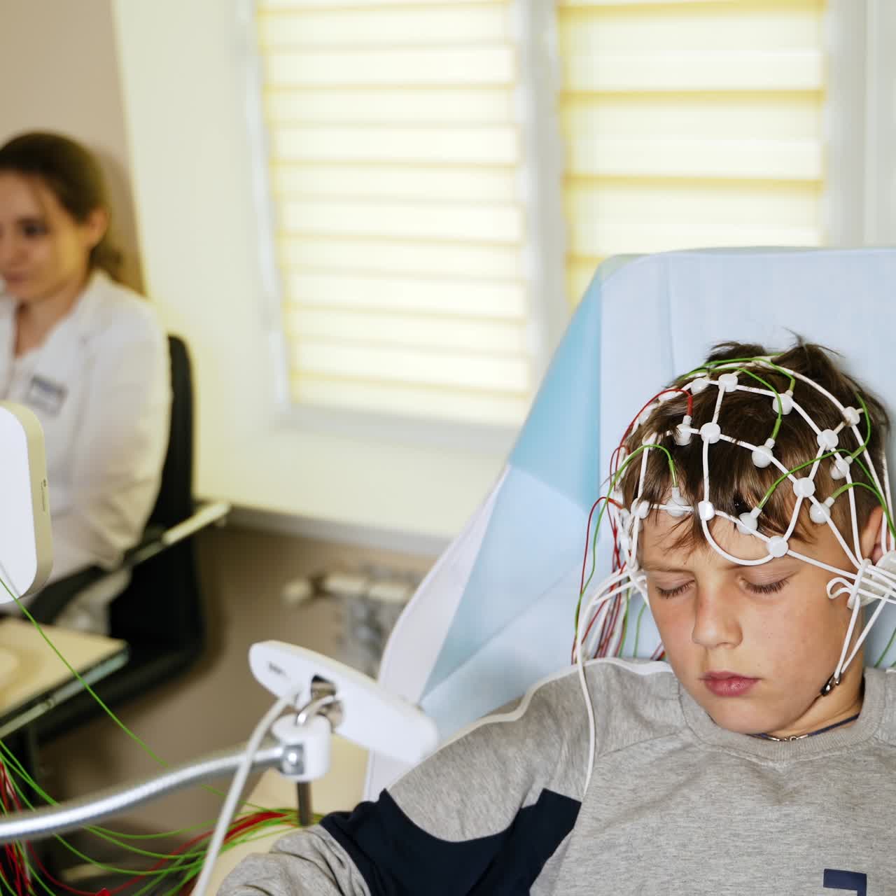 Teenage boy undergoing EEG examination in the hospital. Boy's head connected to lots of sensors. Laboratory medic at backdrop