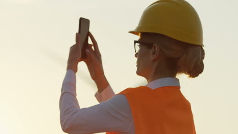 vista lateral de una trabajadora caucásica usando un casco tomando una foto con su teléfono inteligente de las turbinas de los molinos de viento girando al atardecer