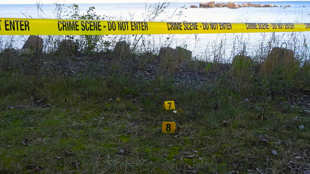 Police line or yellow forensic investigation Crime Scene tape hanging over yellow crime scene police markers on grassy beach shoreline next to bullets and bitcoin money coins as evidence, ocean water