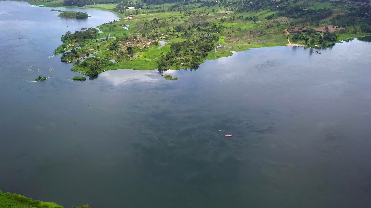 Aerial view of red boat carrying passengers on River Nile in Uganda, surrounded by dark waters, green riverbanks, and scattered shoreline settlements, setting for recreation, travel, and exploration