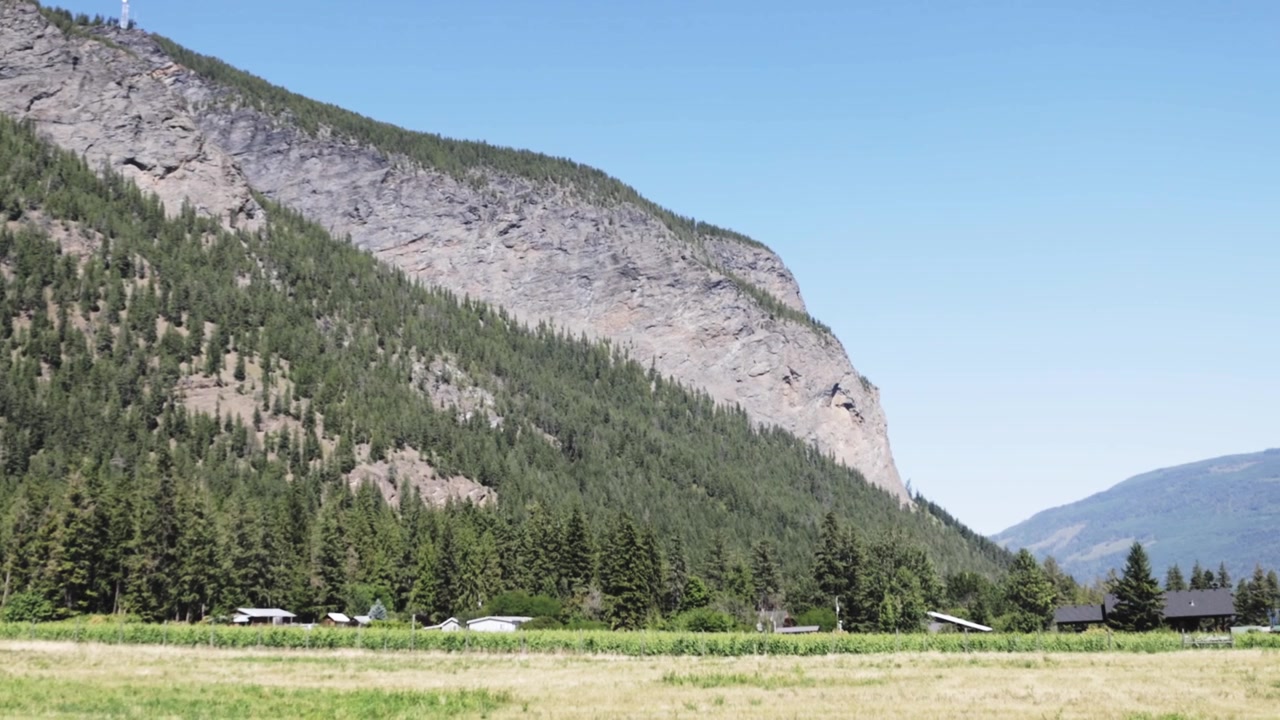 The camera pans from Bastion Mountain’s forested slope toward an open Sunnybrae farm field, showing scattered buildings, trees, and distant ranges under clear daylight in this BC landscape.