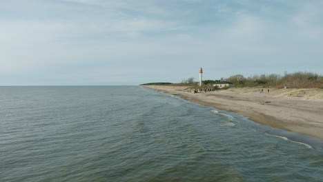 aerial establishing view of white colored pape lighthouse, baltic sea coastline, latvia, white sand beach, large waves crashing, sunny day with clouds, wide drone shot moving froward ascending