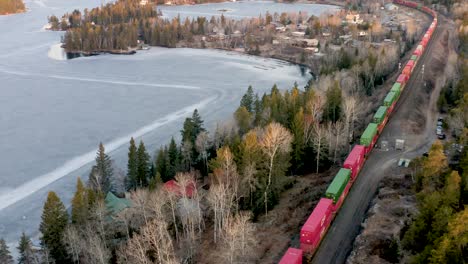 aerial follow cam of a colorful freight train passing by small group of cottages in the canadian shield