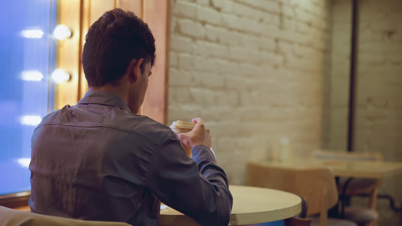 Student in formal shirt sits alone at table inside cozy cafe, holding coffee cup and looking down with thoughtful expression, surrounded by water bottle, smartphone, tissue holder, backpack, and warm window lighting