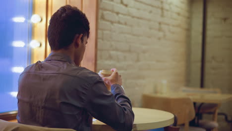 student in formal shirt sits alone at table inside cozy cafe, holding coffee cup and looking down with thoughtful expression, surrounded by water bottle, smartphone, tissue holder, backpack, and warm window lighting