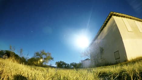 Lapso-De-Noche---Cielo-Azul-Con-La-Luna-Y-Las-Estrellas-Volando-Por-Un-Campo-Con-Un-Viejo-Edificio-Abandonado