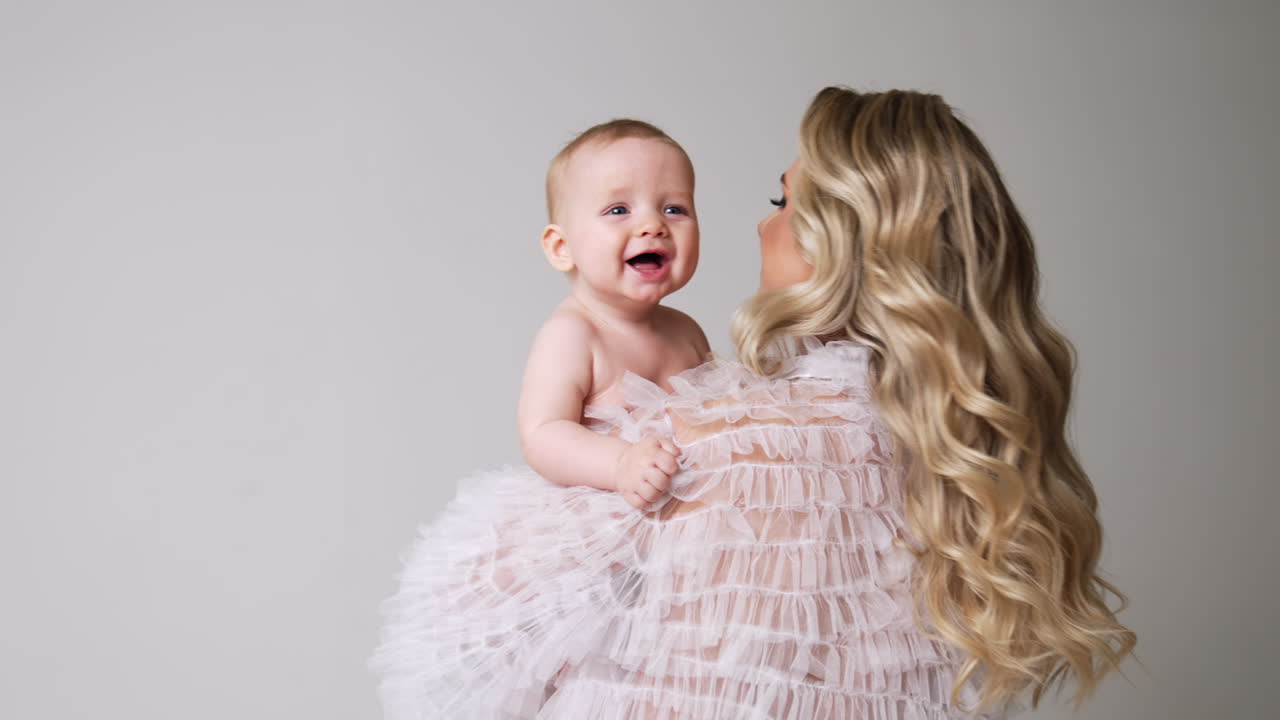 Adorable Caucasian baby smiling sweetly to camera. Naked child in mom's hands at white backdrop. Mother circling with her child.
