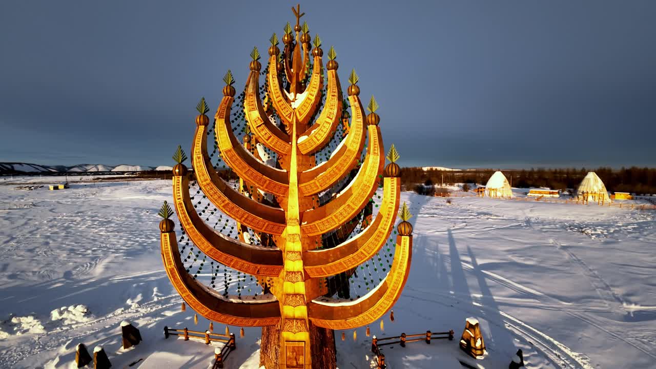 En gyllene, trädliknande struktur står på ett snötäckt fält under en mörk himmel, omgiven av små hyddor.