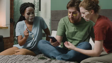 three rommates with facial mask watching smartphone sitting on the bed in bedroom