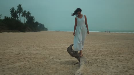 Frau-Balanciert-In-Der-Abenddämmerung-Auf-Einem-Baumstamm-Am-Strand,-Palmen-Im-Hintergrund,-Lässige-Sommerstimmung