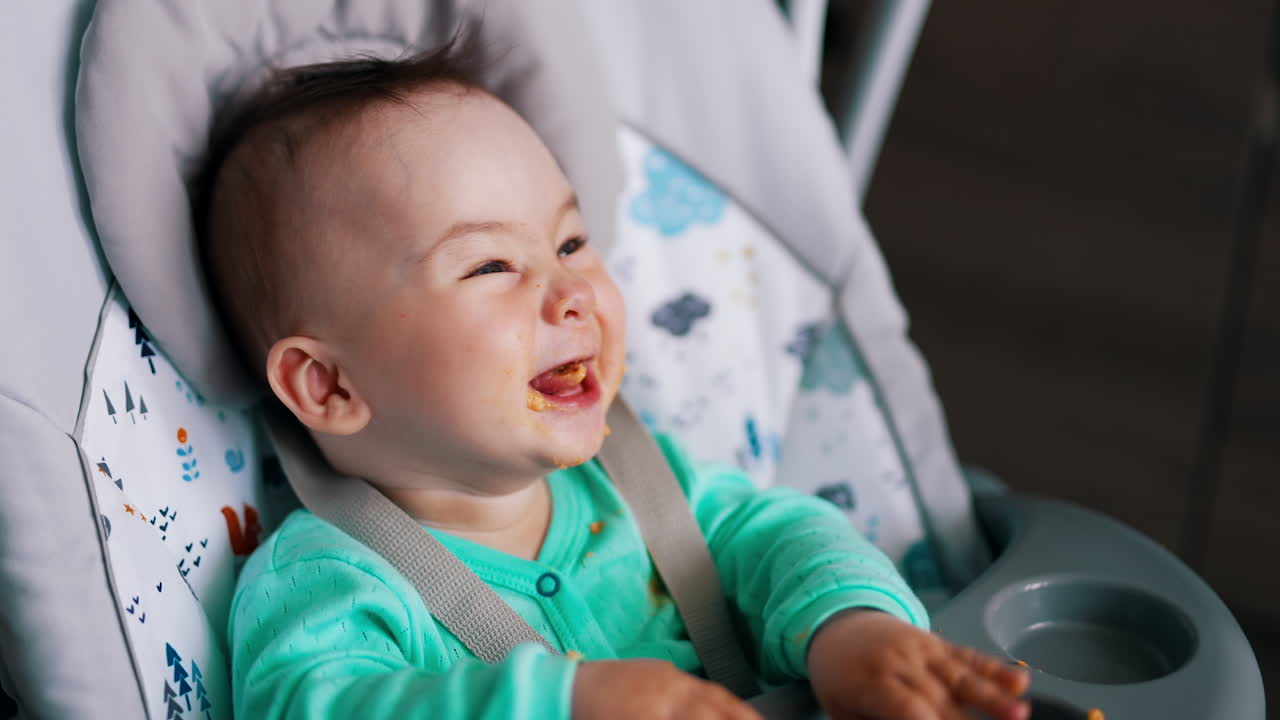 Funny kid sitting in a baby chair is fed from spoon. Childs waves hands and smiles. Close up.