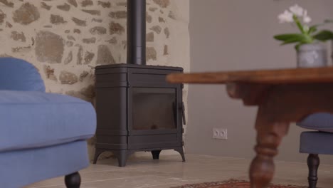 en una pequeña sala de estar, una estufa de leña de metal negro en la esquina de la pared de piedra, una mesa de café de madera con sillas azules en el primer plano borroso