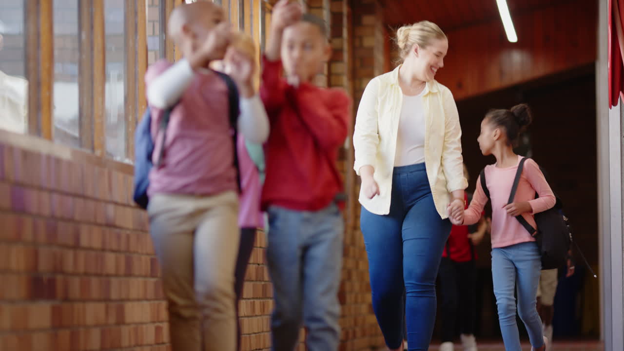 In school hallway, female teacher holding hands with students, smiling and walking together