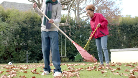 Happy african american mother and son in scarf raking up leaves on lawn in garden, slow motion
