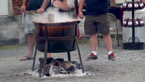 two men standing as one of them stirs steaming apple butter in a large wood fired cauldron outside