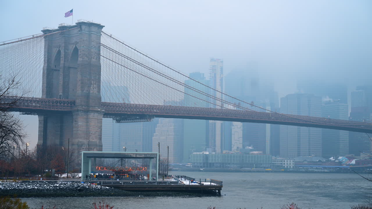 Brooklyn Bridge stone tower and glass carousel in fog. View of bridge tower and Jane's Carousel enclosure on a foggy winter day with snow on waterfront rocks