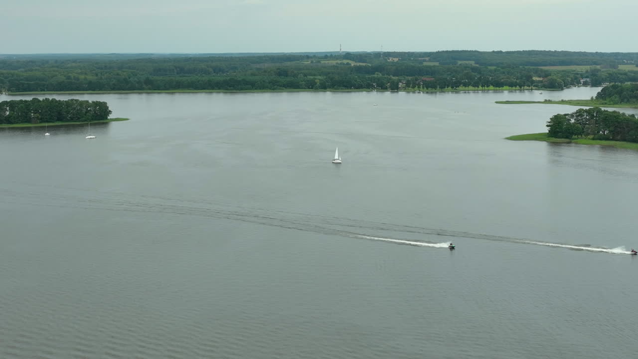 Wide aerial view of two boats leaving trails on the water near a small island, with a sailboat in the distance