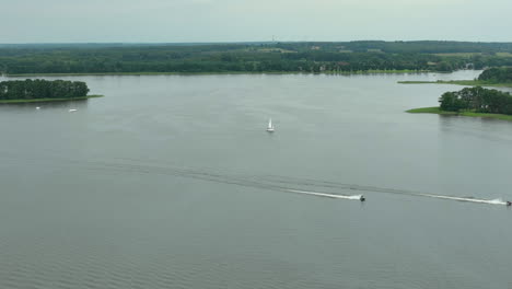 wide aerial view of two boats leaving trails on the water near a small island, with a sailboat in the distance