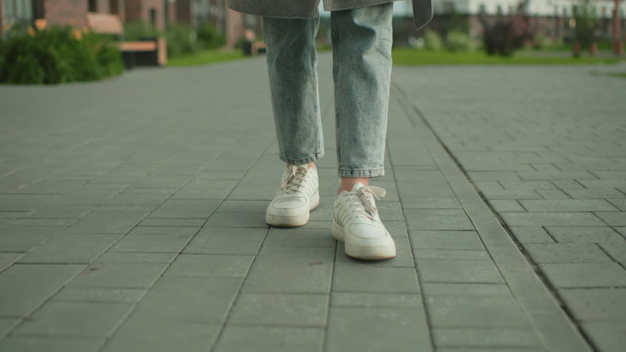 Lower angle close up of person in casual jeans and sneakers walking backwards on paved pathway outdoors, urban street scene in residential environment