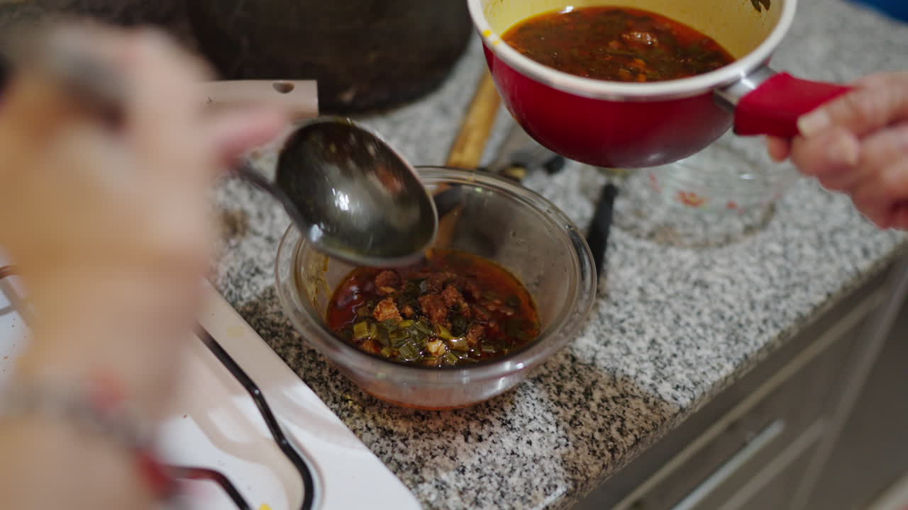 Cook ladles hot locro sauce made from sauteed onions, red and green peppers, rendered chorizo fat and paprika into glass bowl on a granite countertop, the rich orange-red liquid shimmering slow motion