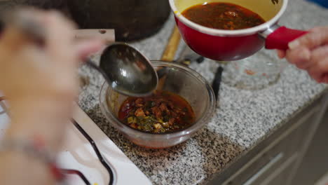 cook ladles hot locro sauce made from sauteed onions, red and green peppers, rendered chorizo fat and paprika into glass bowl on a granite countertop, the rich orange-red liquid shimmering slow motion