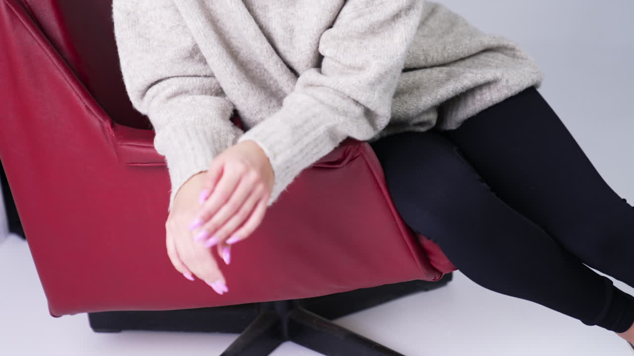 Beautiful smiling brunette girl in warm sweater sitting in red armchair. Model posing in front of camera in studio. White backdrop.
