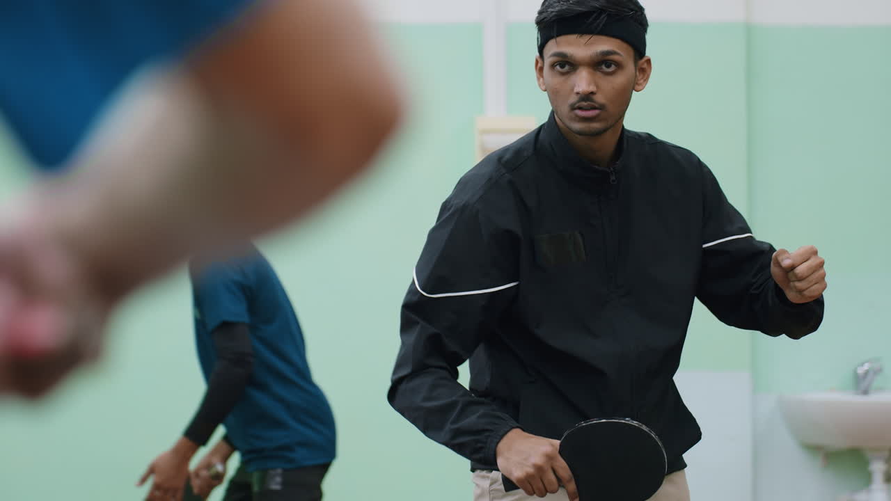 young athlete in black sportswear holding paddle ready for tennis match indoors, intense concentration on face, another player visible in background wearing blue shirt