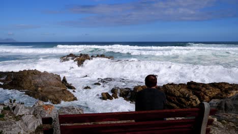 Hombre-Pensativo-En-Un-Banco-Con-Vistas-Al-Océano-Y-Las-Olas-Rompiendo-En-La-Costa-Rocosa