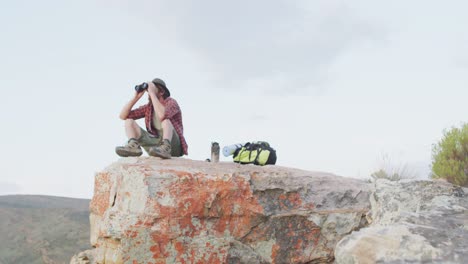 caucasian male survivalist using binoculars, sitting on mountain peak in wilderness