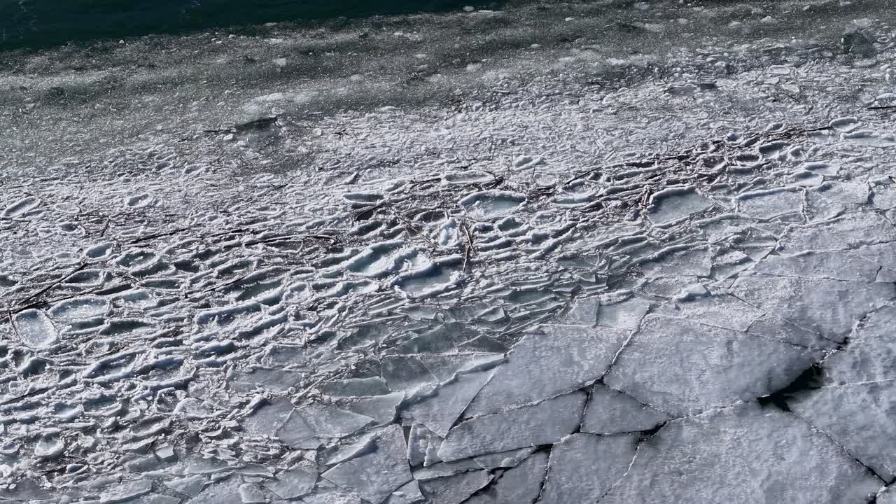 Drone footage captures an overhead view of a partially frozen river with fractured ice plates forming intricate patterns near Whitefish in the Rocky Mountains of Montana