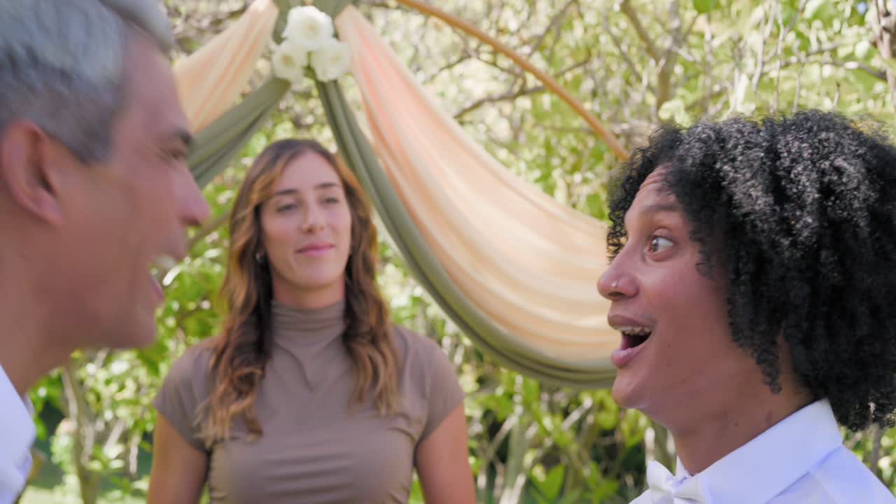 Vertical video: Two men standing at draped arch, left groom speaking vows, officiant holding tablet