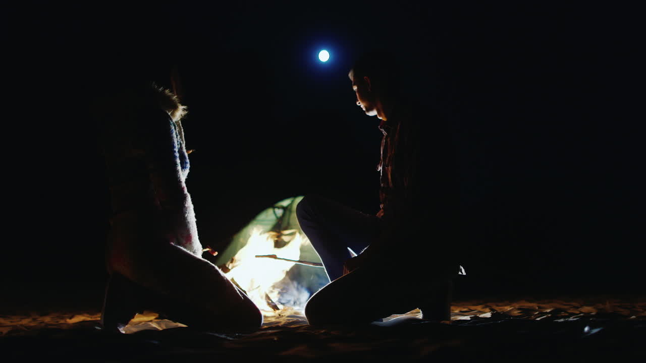 pareja joven relajándose junto al fuego asando salchichas sobre un fuego en el cielo la luna brilla de pie