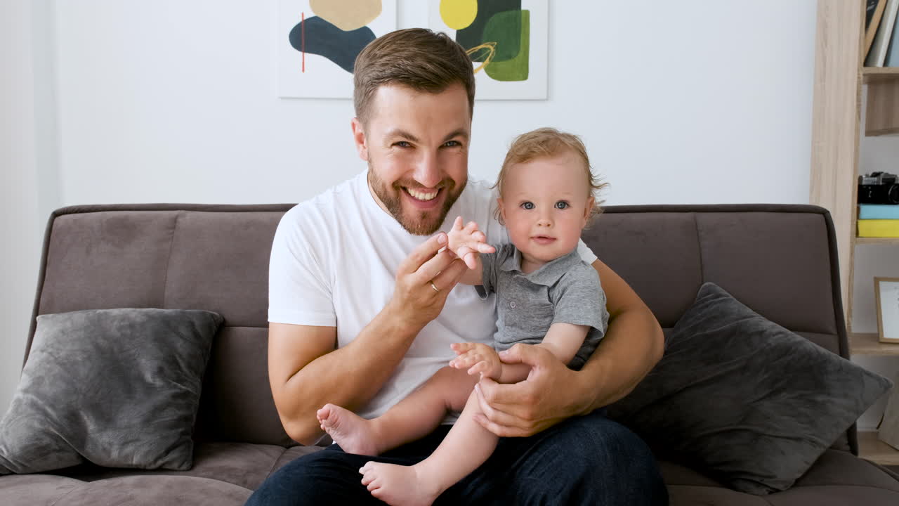 hombre guapo feliz sentado en el sofá en la sala de estar mirando a la cámara durante una videollamada mientras abraza a su lindo niño