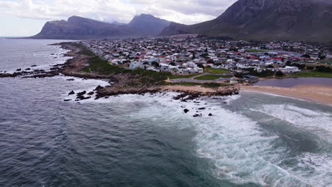 aerial tilt-up over ocean reveals seaside town of kleinmond backed by mountains