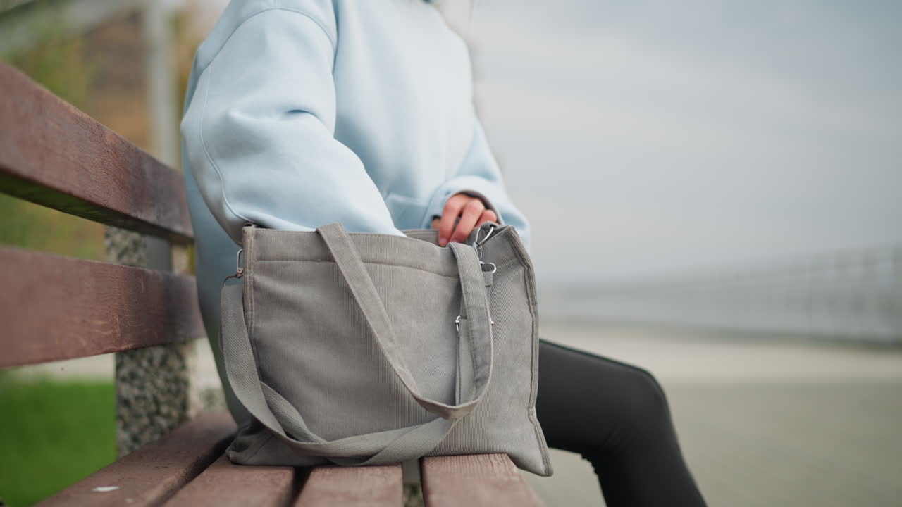 Partial view of woman sitting on concrete bench, searching her bag and pulling out water bottle, peaceful park setting, ideal for relaxation, hydration, and nature breaks