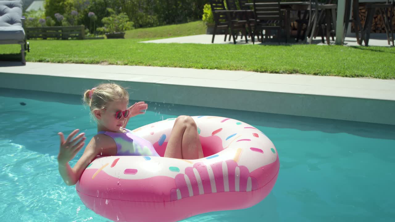 Child girl age 4-6 leaning back then paddling, steering pink donut float at backyard pool edge