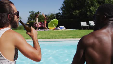 two male diverse friends toasting and drinking beer while sitting by the pool