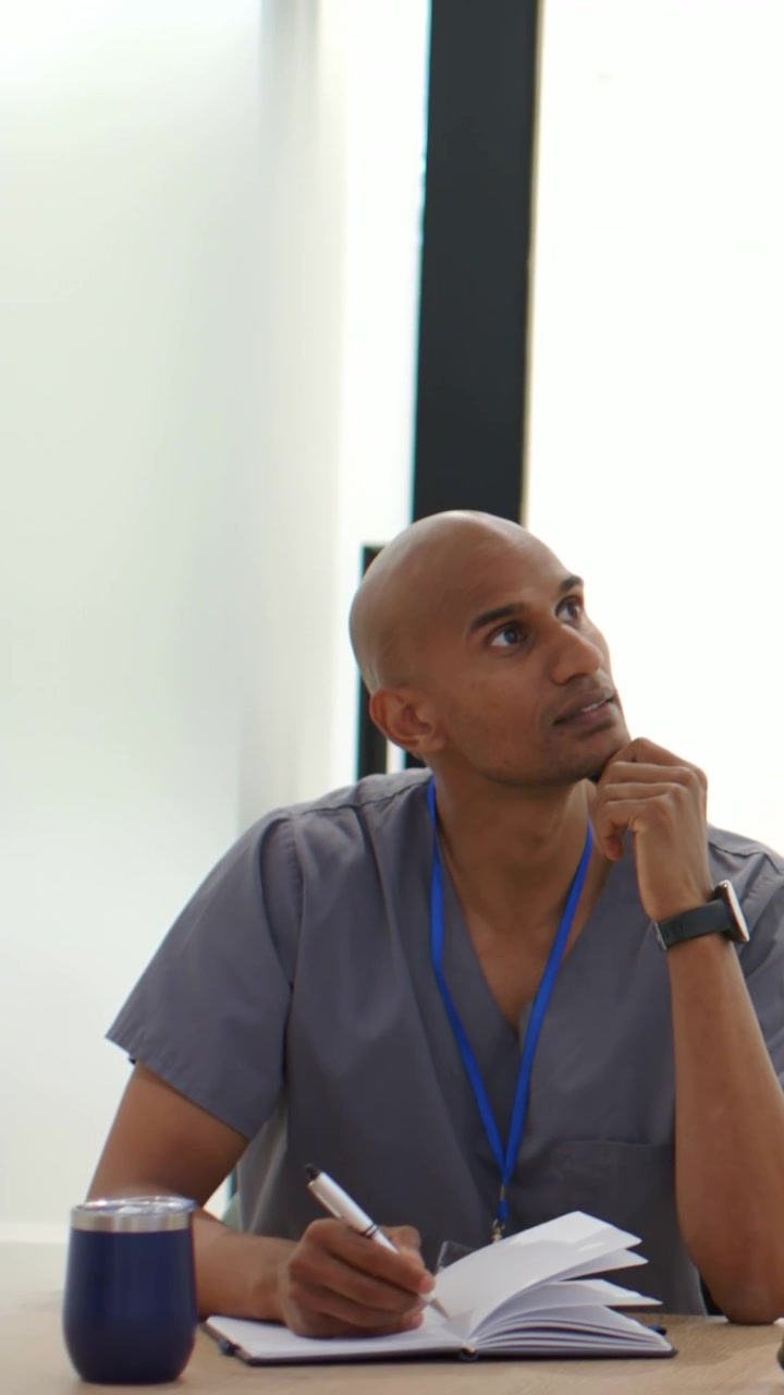 Vertical video: Writing male nurse in scrubs jotting notes at meeting after comment, notebook