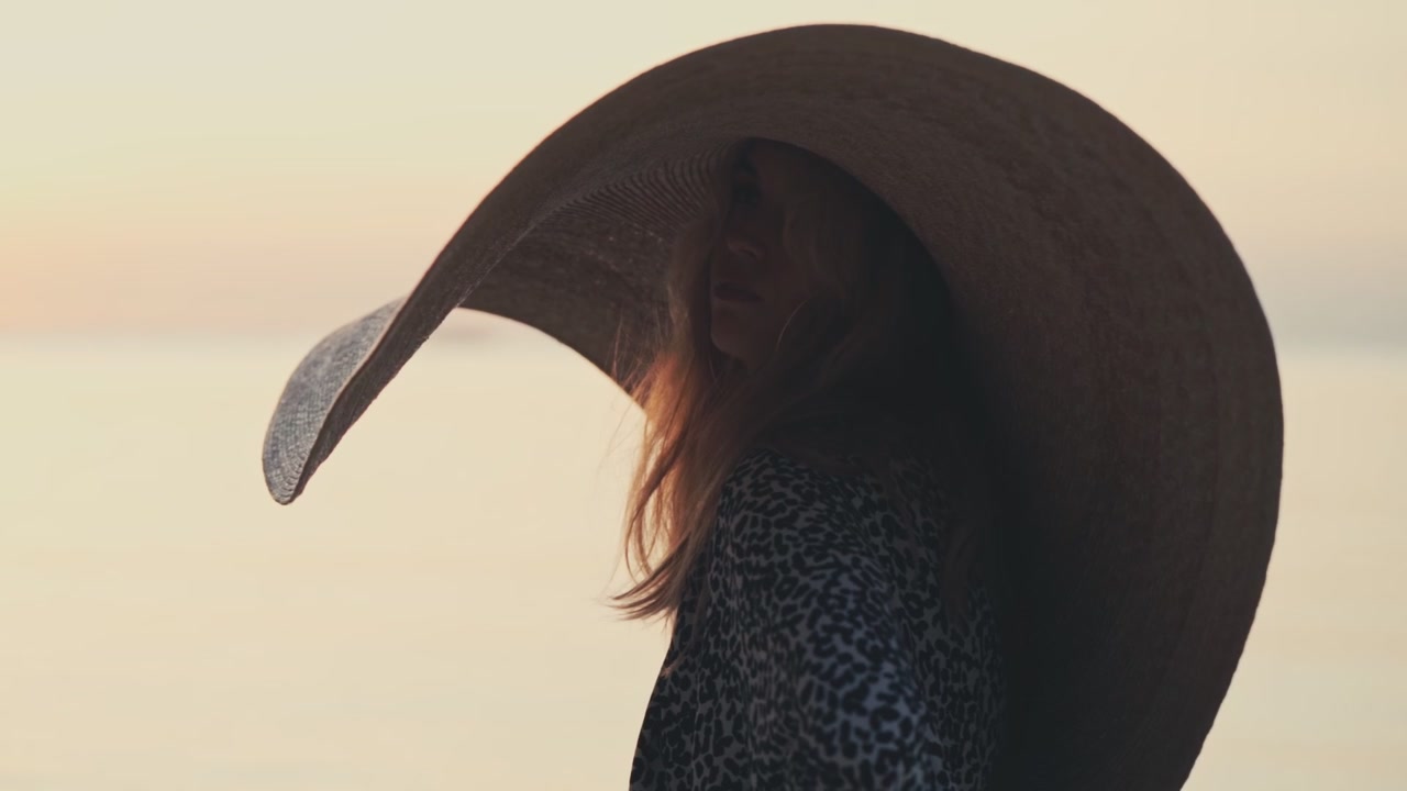 Donna con cappello da sole oversize in piedi in riva al mare al tramonto.