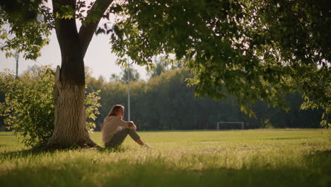 woman sits under tree with arms wrapped around legs, gazing into distance with thoughtful expression, sunlight filters through leaves , with distant trees, and goalpost in background