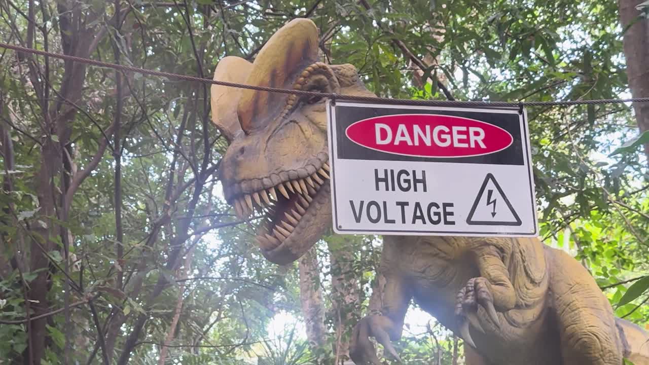 A robotic dinosaur moves behind a high voltage warning sign in a dense forest setting