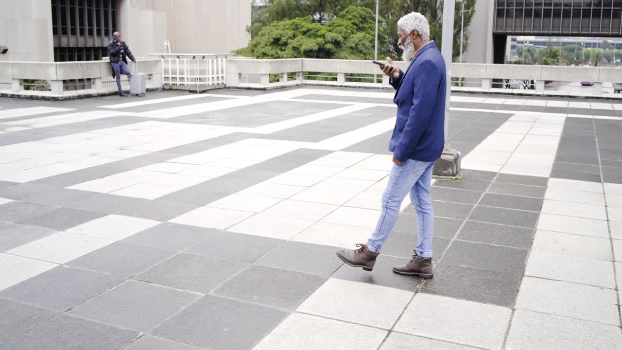 Senior African American man in navy blazer glancing at phone crossing roof plaza checking messages
