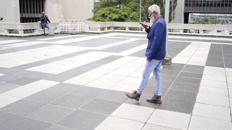 Senior African American man in navy blazer glancing at phone crossing roof plaza checking messages