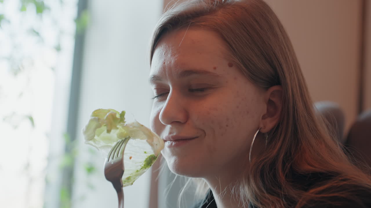 Mujer joven oliendo una hoja de lechuga fresca, un momento de alimentación consciente en una cafetería soleada, retrato íntimo de una bloguera de comida saboreando la textura y el aroma con expresión tranquila, piel pecosa y aretes de aro, suave