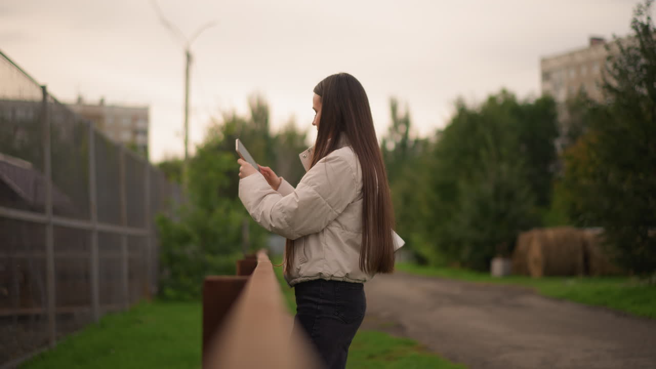 Jovem enviando mensagens perto de uma cerca ao ar livre, caminho suburbano com prédios de apartamentos e árvores, céu nublado, jaqueta acolchoada e cabelo trançado, estudante casual checando mensagens enquanto se apoia no corrimão, suave
