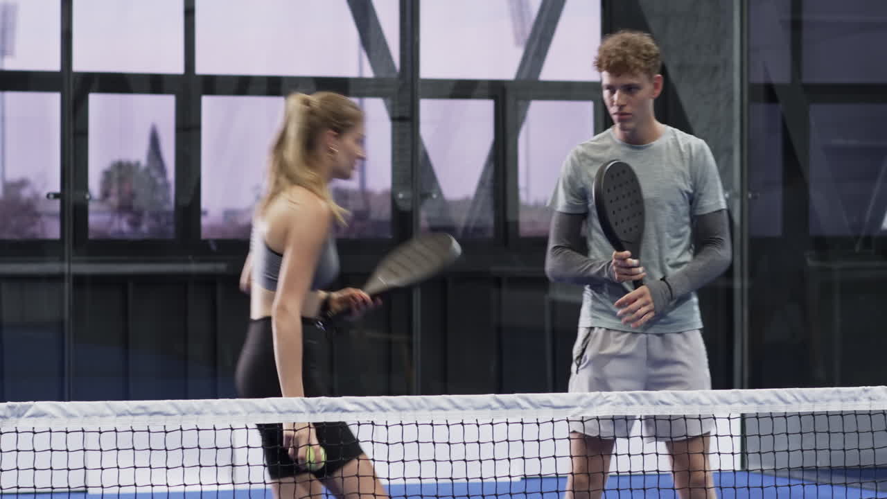 Efter att ha bytt racketar övar vänner forehand och serve vid nätet på en övertäckt bana med boll.