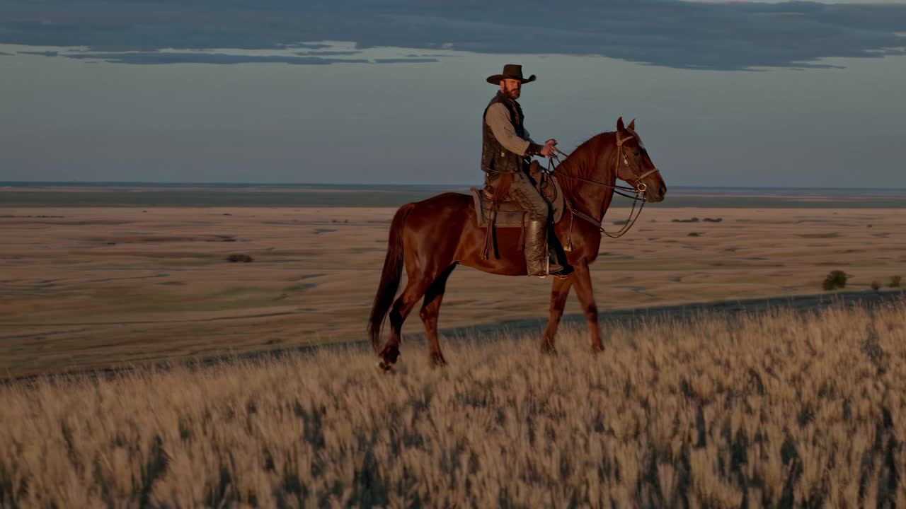 A cowboy on horseback rides through a vast prairie at sunset